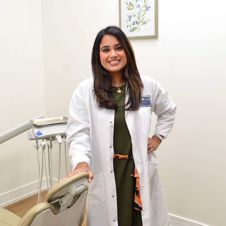 Smiling dentist in white coat standing beside dental chair inside operatory at Smile Circle Dental